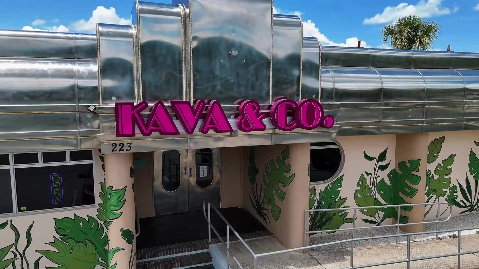 The Kava and Company Jacksonville Beach building at 223 9th Ave S, chrome awnings and a pink neon sign above monstera murals